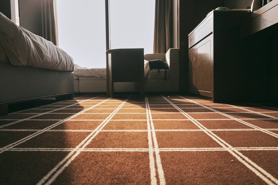 A close-up view of a carpeted floor in a residential bedroom, featuring a brown carpet with a white grid pattern. The room is well-lit by natural sunlight coming through a large window with beige curtains. Visible furnishings include a white bed with a slightly rumpled comforter on the left, a dark-colored chair and a side table positioned near the window, and a wooden dresser on the right. The carpet appears clean and well-maintained, enhancing the room's tidy and hygienic appearance, consistent with surface cleaning and deep cleaning practices discussed on ilfordcarpetcleaning.com for avoiding hidden charges in Ilford. The lighting highlights the fresh, dust-free state of the flooring, emphasizing the importance of regular sanitisation for a spotless and inviting domestic space.