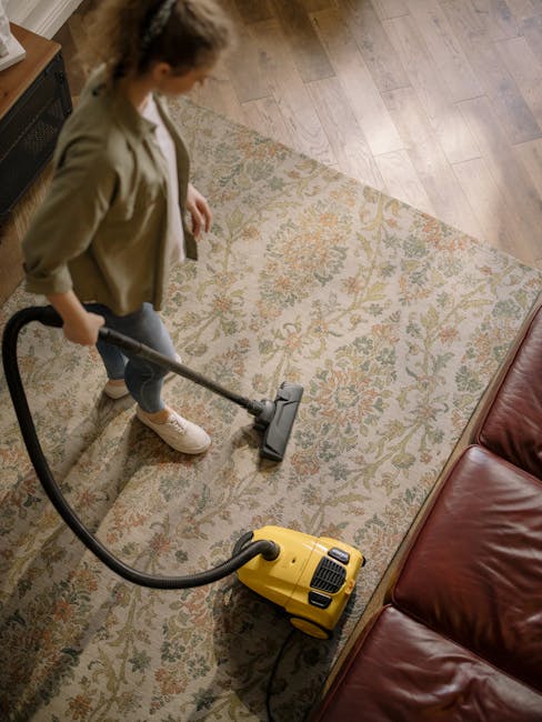 A person operating a yellow vacuum cleaner with a black hose and a wide cleaning head, removing dust and dirt from a patterned carpet in a living room. The carpet features an intricate floral design with shades of beige, orange, and green, set on a wooden floor with visible planks. The room is illuminated by natural light, highlighting the clean and well-maintained surface. To the right, there is a red leather sofa, and in the background, part of a wooden side table or cabinet can be seen. This scene exemplifies domestic surface cleaning, and it illustrates the professional cleaning services provided by Ilford Carpet Cleaning, emphasizing thorough deep cleaning and hygience maintenance.