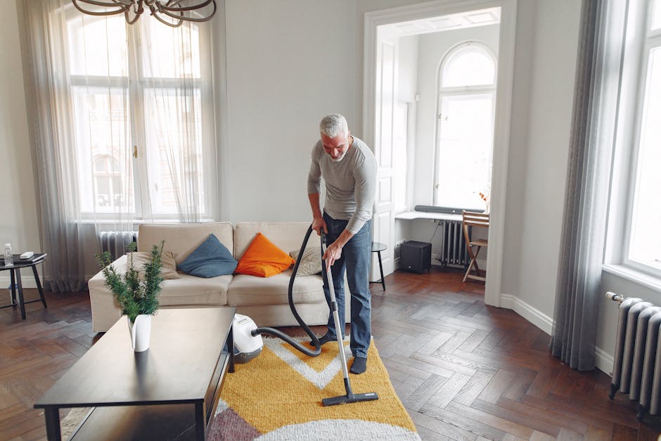 An elderly man with gray hair and a beard is performing surface cleaning of a living room with a vacuum cleaner. The room features a cream-colored sofa adorned with a blue and an orange cushion, positioned against large windows with sheer curtains allowing natural daylight. The dark wooden flooring is partially covered by a yellow patterned rug. A small black side table with decorative items, including a plant in a white vase, is situated next to the sofa. In the background, a small desk and chair are visible near another window, with a radiator beneath it. The room appears neat and well-maintained, with bright natural lighting emphasizing the clean surfaces. Ilford Carpet Cleaning offers expert cleaning services to ensure homes like this remain hygienic and polished, ideal for deep cleaning and maintaining domestic hygiene standards.