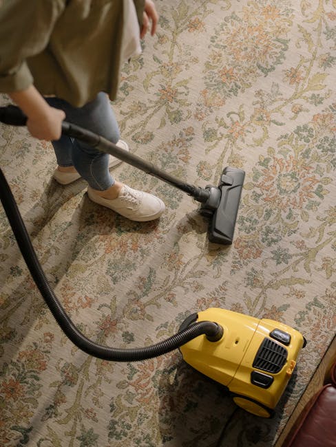 A person using a vacuum cleaner with a yellow body and black hose on an ornate, traditional patterned carpet in a residential room. The individual, dressed in casual clothing and white sneakers, is holding the vacuum's long handle, applying deep cleaning to the beige and green floral design rug, which appears slightly damp or recently cleaned. The room has warm lighting, and part of a wooden furniture piece can be seen near the carpet. This scene depicts routine surface cleaning and maintenance by Ilford Carpet Cleaning, focusing on thorough domestic or commercial carpet sanitisation.