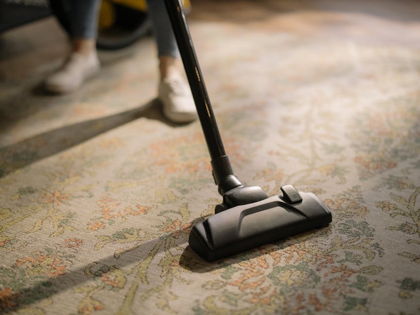 Close-up image of a vacuum cleaner head actively cleaning a patterned area rug with intricate floral motifs in a well-lit room. The vacuum is black with a slightly curved design, and the handle extends upward, held by a person wearing white shoes and dark pants. The surface of the rug appears to be free of visible dust or dirt, indicating a thorough surface cleaning process. Behind the vacuum, part of a person's leg and foot are visible, suggesting an ongoing domestic cleaning session in a residential setting. The image highlights the effectiveness of surface cleaning and deep cleaning techniques used by Ilford Carpet Cleaning for maintaining hygiene and cleanliness in homes.