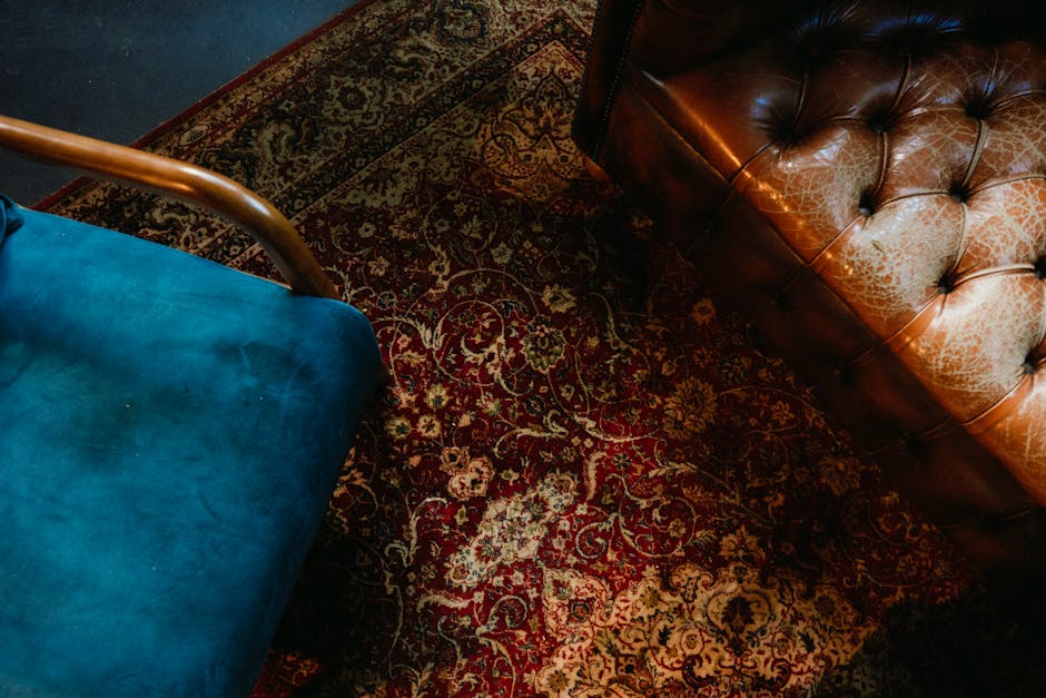 A close-up view of a traditional patterned carpet with intricate floral designs and a red, beige, and dark brown color scheme, situated in a room with wooden furniture and leather upholstery. The carp
