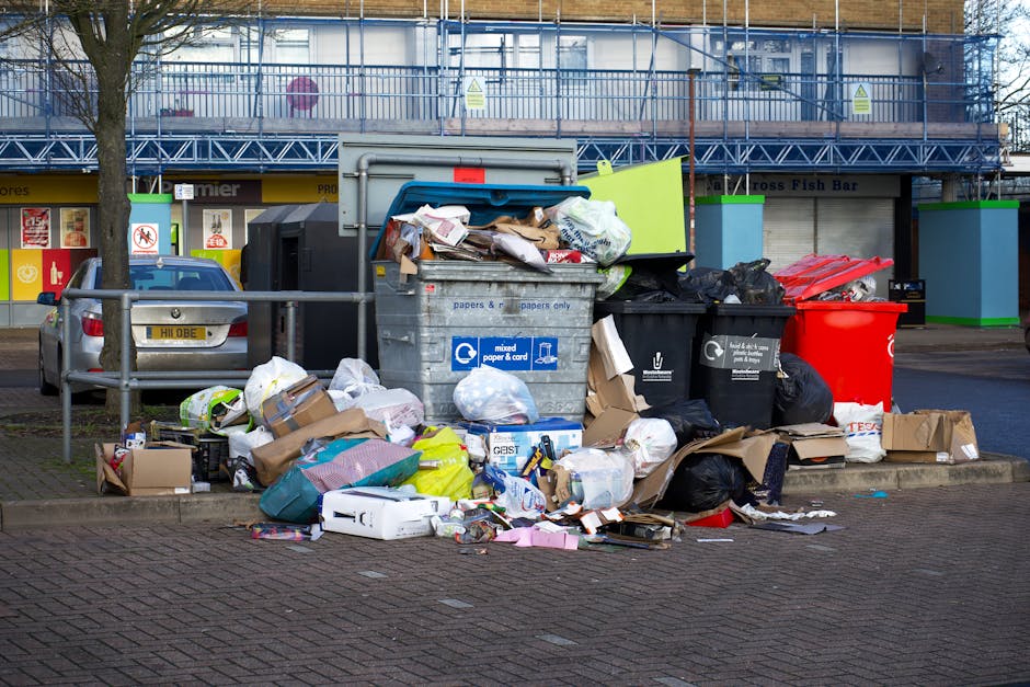 A collection of overflowing waste bins and scattered rubbish on a sidewalk in front of a commercial building with retail shops. The waste includes black, red, and black-and-white patterned garbage bags, flattened cardboard boxes, paper and plastic packaging, and loose discarded items. The bins are situated behind a metal railing, with one larger grey mixed paper and cardboard bin, and smaller black bins. The area appears untidy and in need of cleaning to comply with hygiene standards. Bright daylight illuminates the scene, highlighting the mess and the surrounding urban environment. The image relates to proper waste management and cleaning regulations as outlined by Redbridge Council for residential and commercial cleaning, as provided by Ilford Carpet Cleaning.
