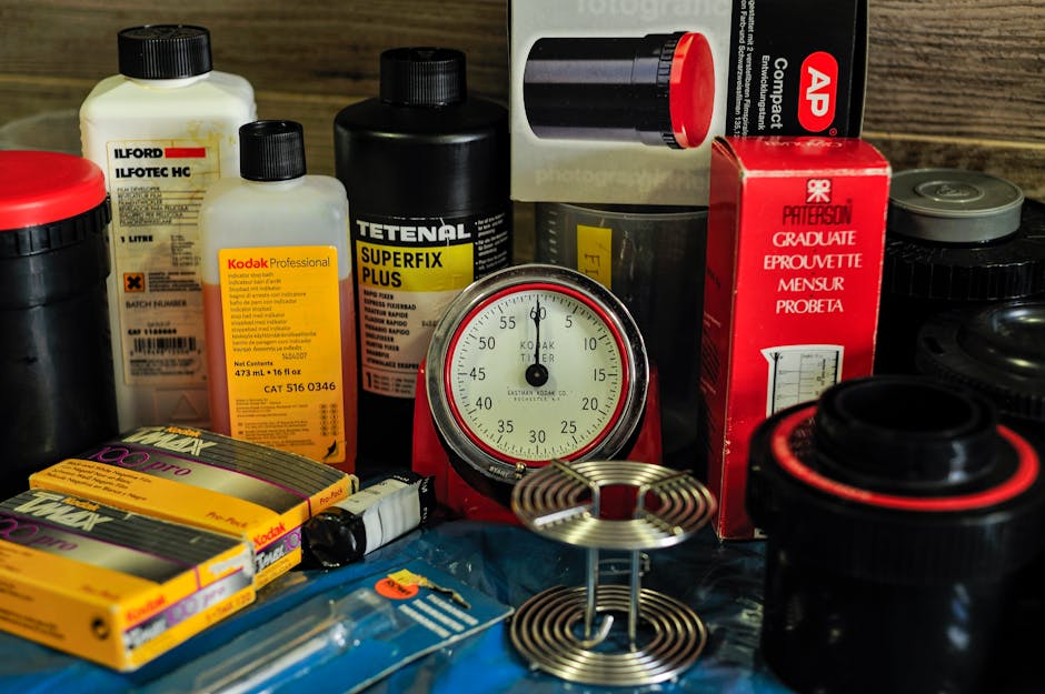A collection of cleaning supplies and tools arranged on a wooden surface, including bottles of chemicals with labels indicating brands such as Kodak Professional and Tetenal Superfix Plus, a red-capped bottle, a black container, and a red and white box, alongside a round metal timer and a blue package with cleaning accessories. The background shows a wooden surface with a subtle grain pattern, and the overall setting suggests a preparation area for surface cleaning or deep cleaning tasks. The scene is well-lit, emphasizing the cleanliness and organization of the various materials used for domestic or commercial sanitisation, reflecting the cleaning procedures aligned with regulations like those from Redbridge Council for waste management. Ilford Carpet Cleaning offers expertise in maintaining hygienic surfaces, helping ensure safe and effective cleaning processes.