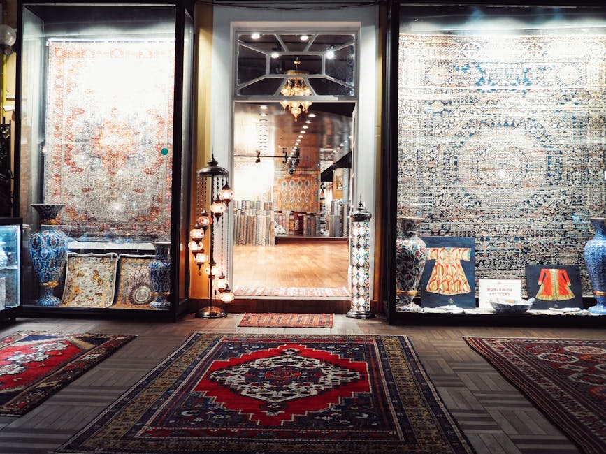 Interior view of a rug shop displaying various patterned rugs and carpets on the walls and floor, with a wooden floor in the background illuminated by warm lighting and a chandelier. The shop features decorative vases and lamps on either side of the entrance, which has glass display windows showcasing additional rugs and ornaments. The space appears clean and well-maintained, emphasizing surface cleanliness and presentation, consistent with professional carpet and rug cleaning standards by Ilford Carpet Cleaning. The overall atmosphere highlights the importance of deep cleaning, sanitisation, and careful surface maintenance of decorative rugs and textiles for domestic or commercial settings.