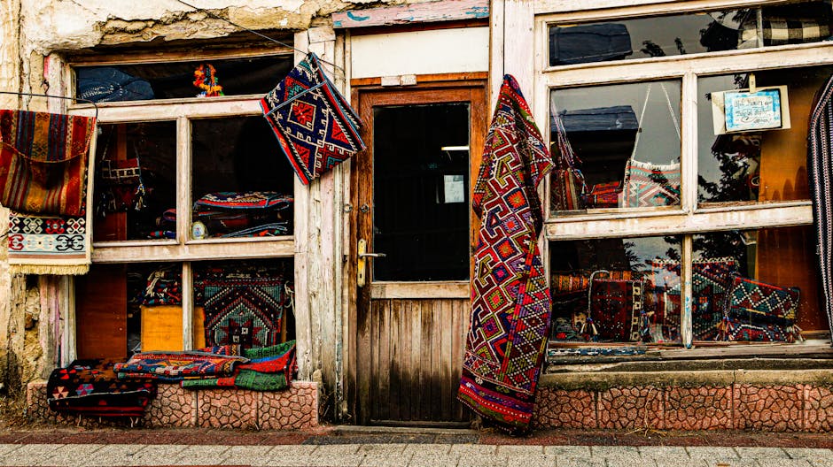The image depicts the exterior of a shop with wooden framing, displaying colorful, patterned rugs hanging from the window and stacked along the ledge. The shop's front has a small glass-paned door, flanked by large glass windows showcasing more rugs and textiles inside. The surfaces include wooden window trim, a concrete sidewalk, and a stone or tile platform at the base of the shopfront. The natural lighting indicates daytime, with reflections of the outside environment visible on the glass. The display appears arranged in a casual manner, highlighting a variety of woven rugs with intricate geometric designs and vibrant colors. The cleanliness of the display is maintained, with the rugs appearing clean and well-organized, aligned with professional cleaning and maintenance practices as promoted by Ilford Carpet Cleaning, especially relevant when considering surface and fabric management in commercial settings.
