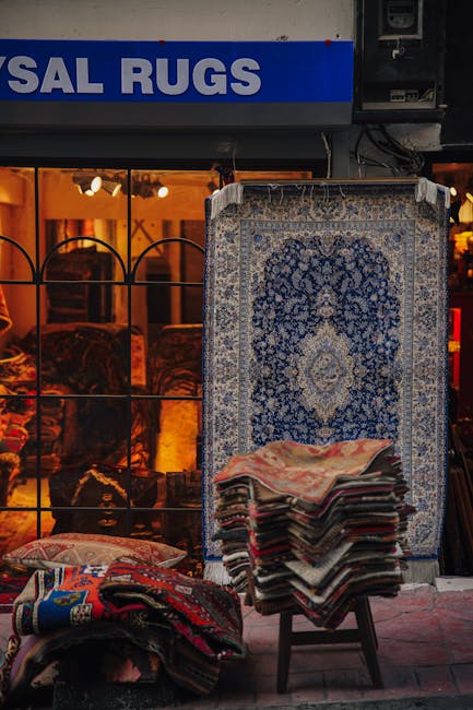 Interior view of a rug shop displaying various patterned rugs and carpets on the walls and floor, with a wooden floor in the background illuminated by warm lighting and a chandelier. The shop features decorative vases and lamps on either side of the entrance, which has glass display windows showcasing additional rugs and ornaments. The space appears clean and well-maintained, emphasizing surface cleanliness and presentation, consistent with professional carpet and rug cleaning standards by Ilford Carpet Cleaning. The overall atmosphere highlights the importance of deep cleaning, sanitisation, and careful surface maintenance of decorative rugs and textiles for domestic or commercial settings.