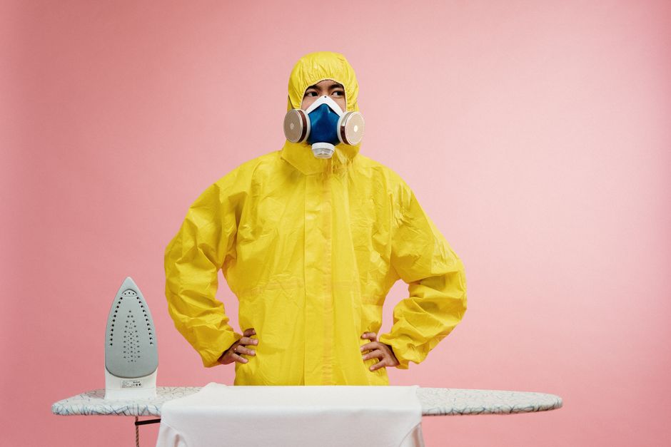 A person dressed in a yellow protective suit with a hood, wearing a blue respirator mask with two filters, stands with hands on hips in a room with a pink wall. In front of them is an ironing board covered with a white cloth, and to the left is a steam iron. The scene suggests preparation for surface cleaning or domestic cleaning activities, emphasizing hygiene and safety measures before professional cleaning services by Ilford Carpet Cleaning arrive. The bright lighting highlights the clean, shiny surface of the ironing board and the detailed texture of the suit and equipment, indicating a focus on thorough sanitation and deep cleaning procedures.