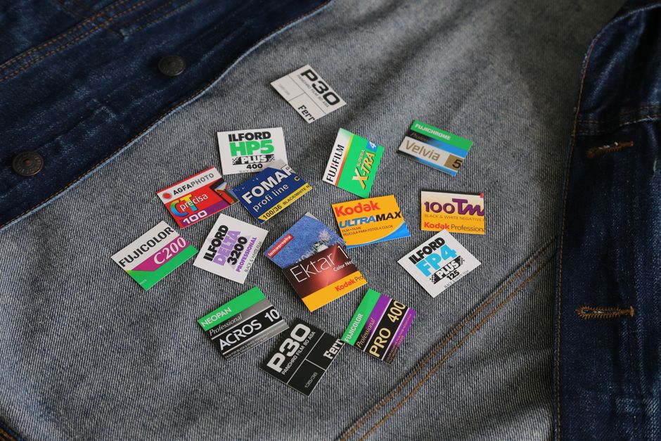 A close-up image of a pair of blue denim jeans laid flat on a surface, with a collection of various photographic film type labels scattered on the front pocket area. The labels are colorful and feature different brand names and specifications such as Ilford HP5 Plus, Kodak UltraMax, Fomapan Profi Line, FujiColor, and others, indicating different film types and ISO ratings. The denim appears clean and slightly textured, with visible stitching and metal rivets; the lighting is even, emphasizing the contrasts of the colorful labels against the dark blue fabric. This image relates to the topic of surface cleaning and maintaining fabric or material cleanliness, highlighting the importance of proper hygiene and product handling in domestic or commercial environments, which is relevant in the context of timely stain management before professional cleaning by Ilford Carpet Cleaning.