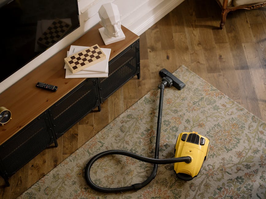 A yellow canister vacuum cleaner with a black hose and metal wand attachment is positioned on a beige, floral-patterned area rug in a living room. The room features wooden flooring, and a black modern TV console with storage drawers is placed against a white wall. On top of the console, there is a white bust sculpture, a closed chessboard, a stack of papers, a remote control, and a gold-colored clock. The area is well-lit, and the scene captures a moment of surface cleaning and general maintenance to ensure hygiene and cleanliness, with Ilford Carpet Cleaning's service focus implied in the context of household or commercial deep cleaning.